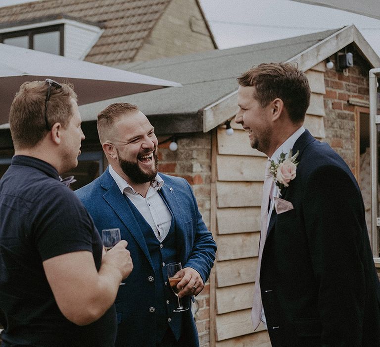 Groom in a navy blue blazer with light pink rose buttonhole flower laughing with his guests at his back garden wedding 