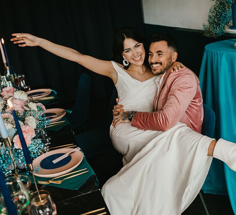 Bride in white boots with block heels sitting on her grooms lap at their pink and teal wedding reception 