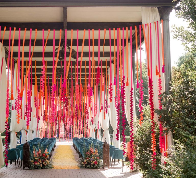 Wedding ceremony venue outdoors surrounded by colourful hangings and pastel floral installations and emerald blue velvet chairs