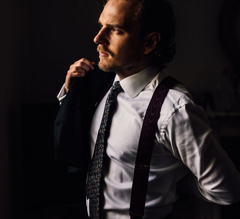 Groom in white shirt, patterned tie and braces puts on jacket by window for Cornish wedding with Halfpenny London Mayfair dress