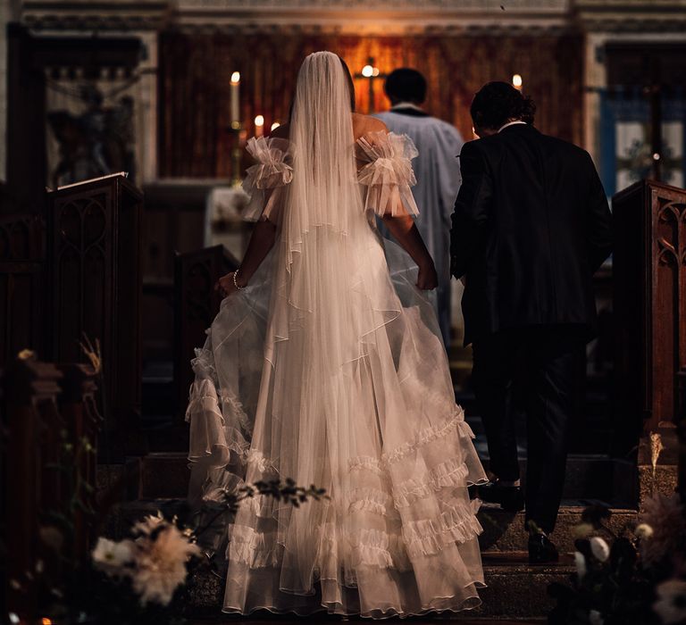 Bride in white Halfpenny London Mayfair dress with ruffle sleeves walks up to the altar with groom in suit during church wedding ceremony in Cornwall