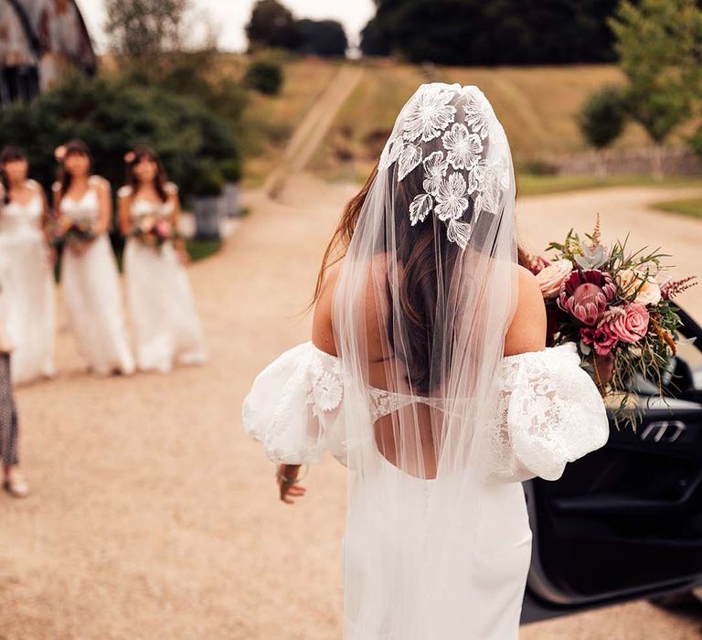 Bridesmaids see bride for the first time as she holds bright floral bouquet and wears lace embellished veil 