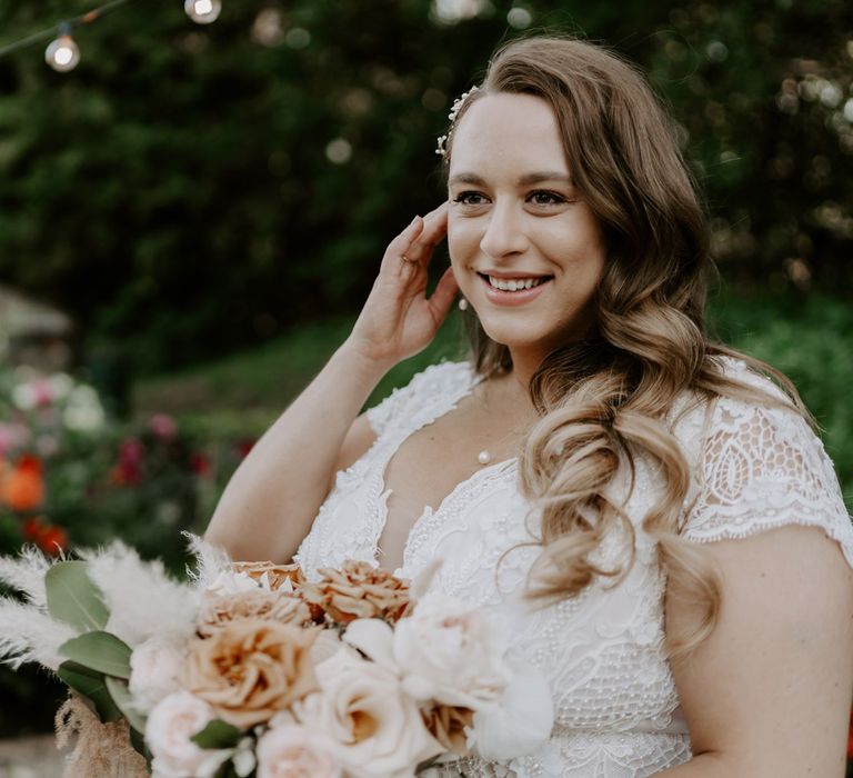 Smiling bride with long curled hair in homemade lace wedding dress and pearl necklace holds mixed rose bridal bouquet as she stands in garden after wedding ceremony