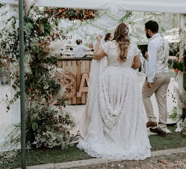 Bride in lace homemade wedding dress with train and open back with curled hair and groom in beige waistcoat and trousers walk into marquee with garlands at outdoor wedding reception