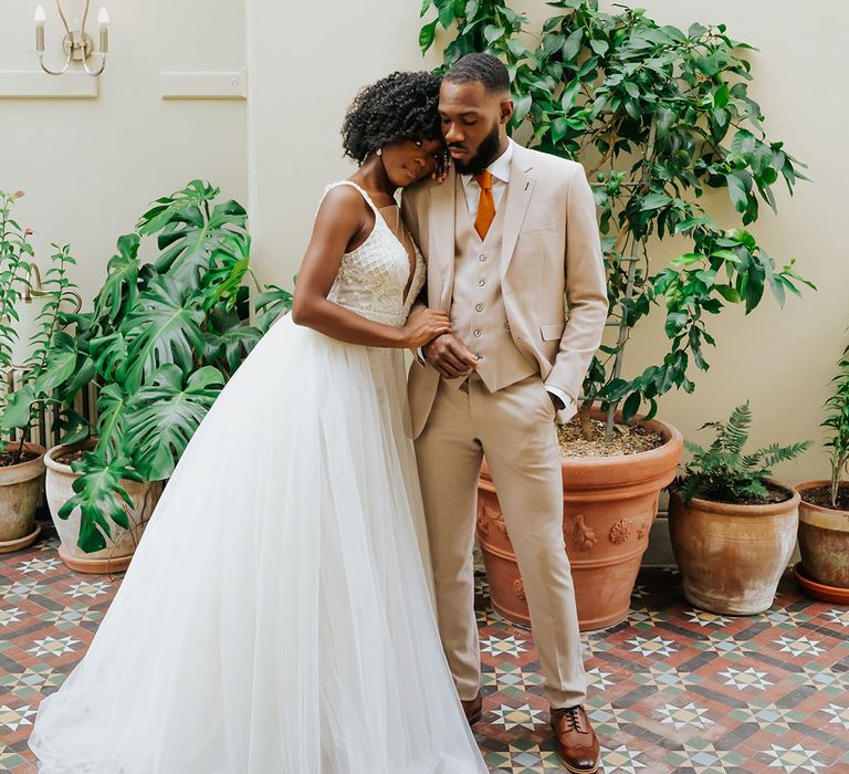 Black bride in a princess wedding dress with tulle skirt embracing her groom in a beige suit with orange tie in a conservatory at Modern Hall, London 