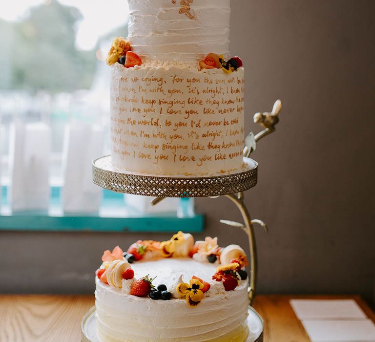 3-tier wedding cake complete with white ruffled frosting, fruit, calligraphy lyrics and gold leaf decor