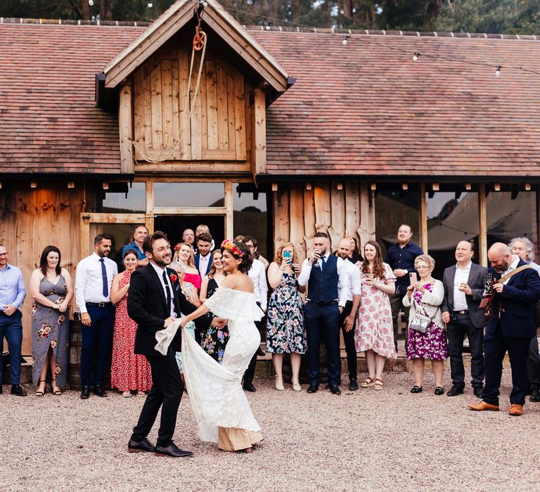 Bride in off the shoulder Grace Loves Lace wedding dress and flower crown dances with groom in black suit in front of wedding guests outside The Bridal Barn in Claverley