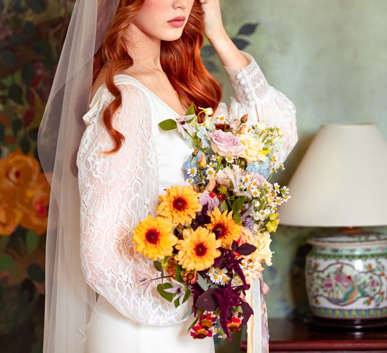 Bride in long lace sleeve wedding dress and Juliet cap veil holds colourful bridal bouquet with daisies and roses