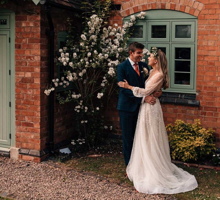 Groom in blue three piece checked suit hugs bride in long mesh sleeve Julie Vino wedding dress and flower crown as they stand outside cottage