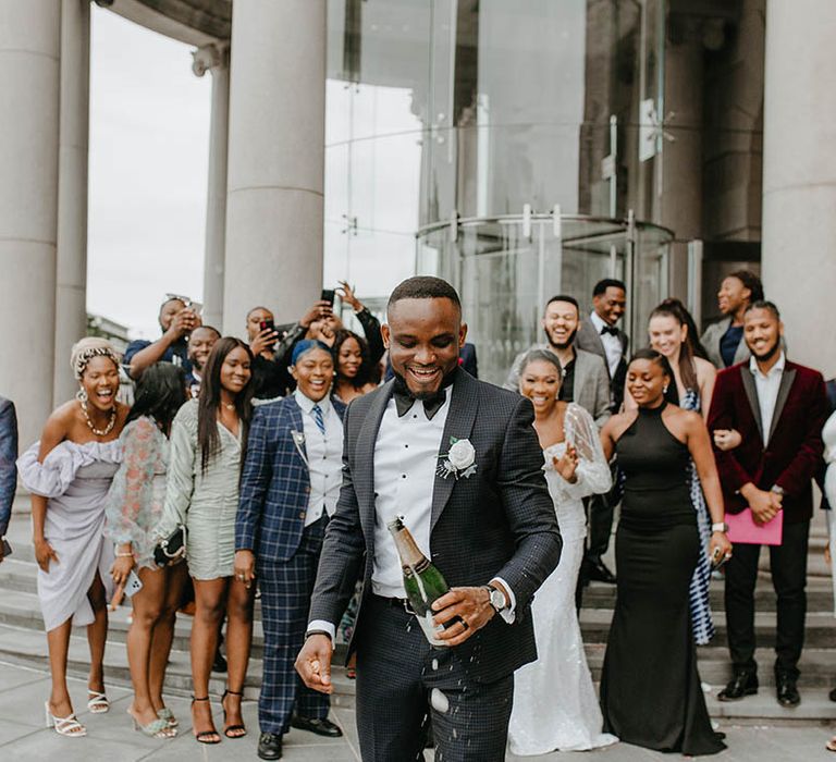 Groom pops champagne outdoors and wedding party stand behind him and celebrate