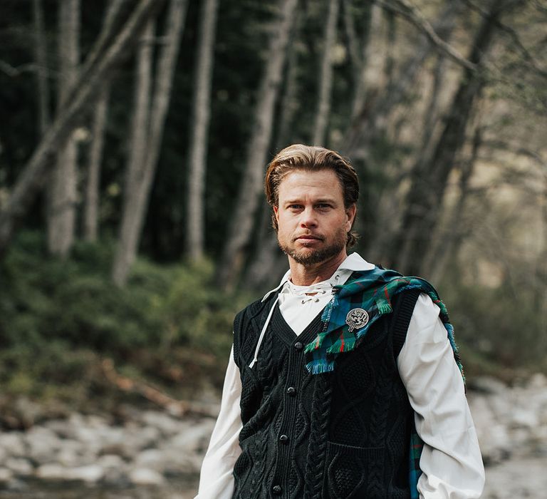 Bearded groom with long wavy hair in tartan kilt and  aran tank top walking through a stream 