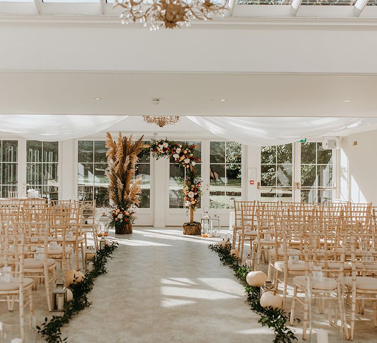 Wedding ceremony aisle complete with white pumpkin decoration, lanterns with white candles and wooden chairs