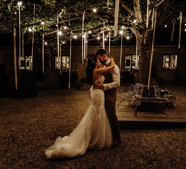 Bride in lace and tulle wedding dress with train dances outside with arms around groom in white shirt and brown tweed waistcoat under tree with bulb lights