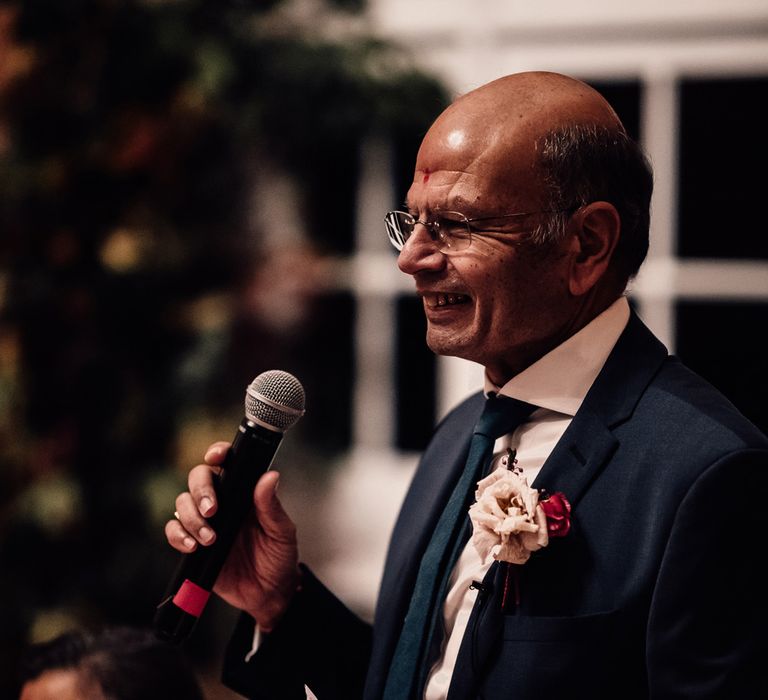 Father of the bride in blue suit with floral buttonhole holds microphone as he makes speech at wedding reception