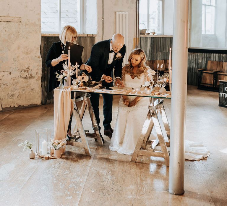 Bride in v neck wedding dress with sleeves sits signing wedding register with groom in navy tweed suit at Holmes Mill wedding