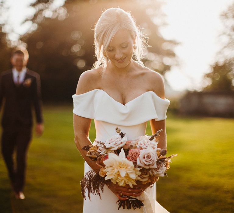 Bride in sweetheart off the shoulder wedding dress holding neutral wedding bouquet as groom approaches her in the background