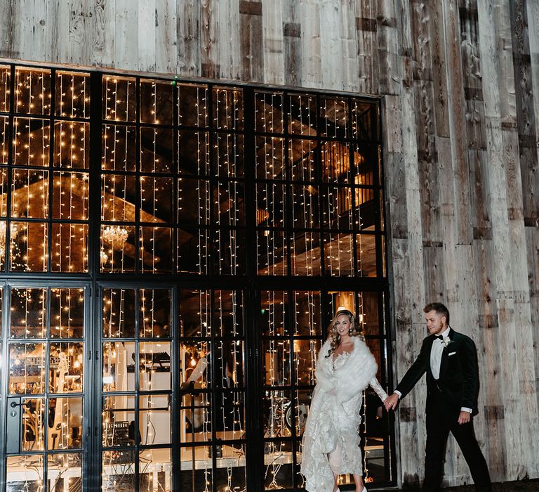 Bride in lace fitted wedding dress with white fur shawl walks with groom in black tie in front of fairy lights wedding