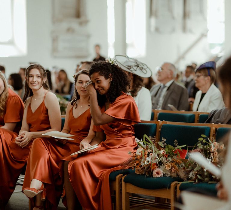 Bridesmaids in orange satin dresses in various styles sit together at church ceremony wedding as one of them cries