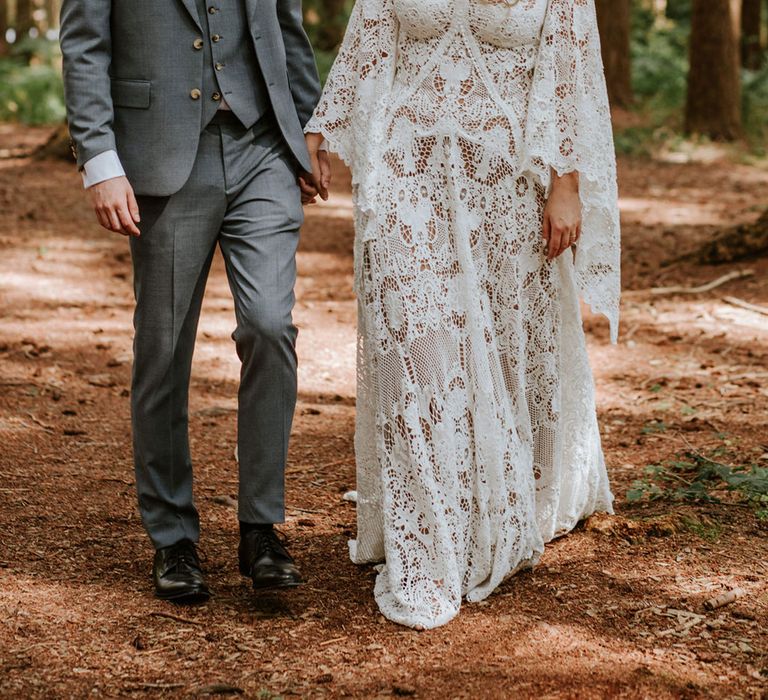 Bride in medieval style lace wedding dress with custom sleeves holding hands with groom in grey suit and pink tie in woodland wedding setting 