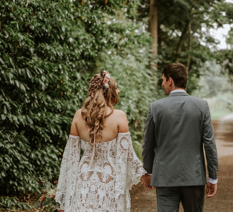 Bride in lace wedding dress with custom sleeves holding hands with groom in grey suit for woodland wedding