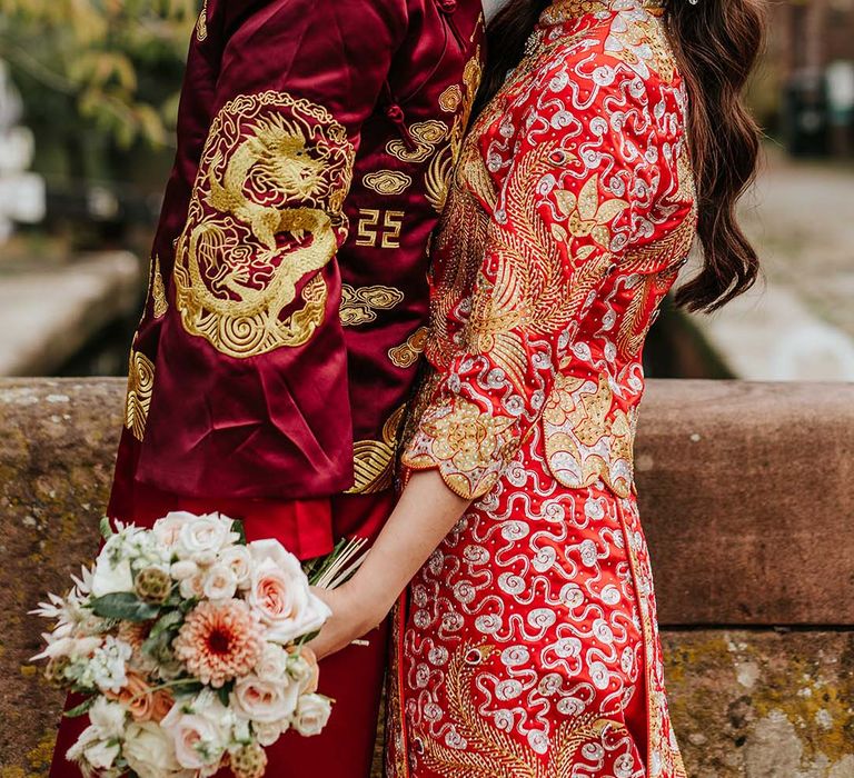 Bride and groom in traditional red and gold Chinese wedding attire with bride wearing pearl and silver accessories holding autumnal bouquet