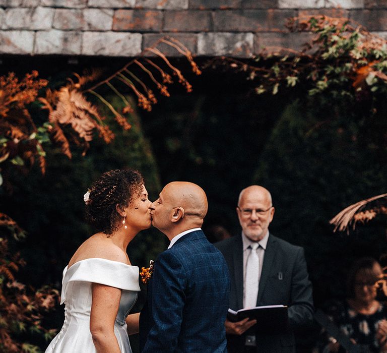 Groom in blue checkered suit kisses bride in off the shoulder wedding dress for outdoor wedding at Askham Hall 