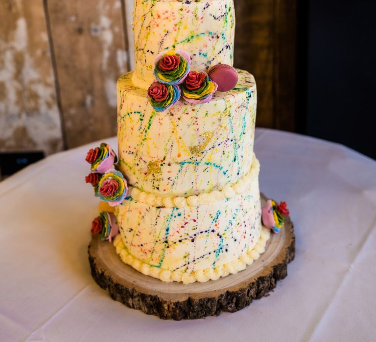 Three tier white frosted rainbow paint splattered wedding cake with rainbow roses, macarons and personalised wooden heart topper