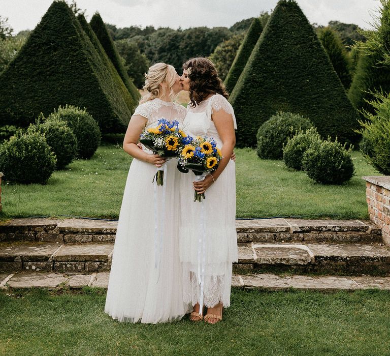 Brides holding sunflower and gypsophila wedding bouquets and sheer high neck wedding dresses share a kiss in the gardens of the venue