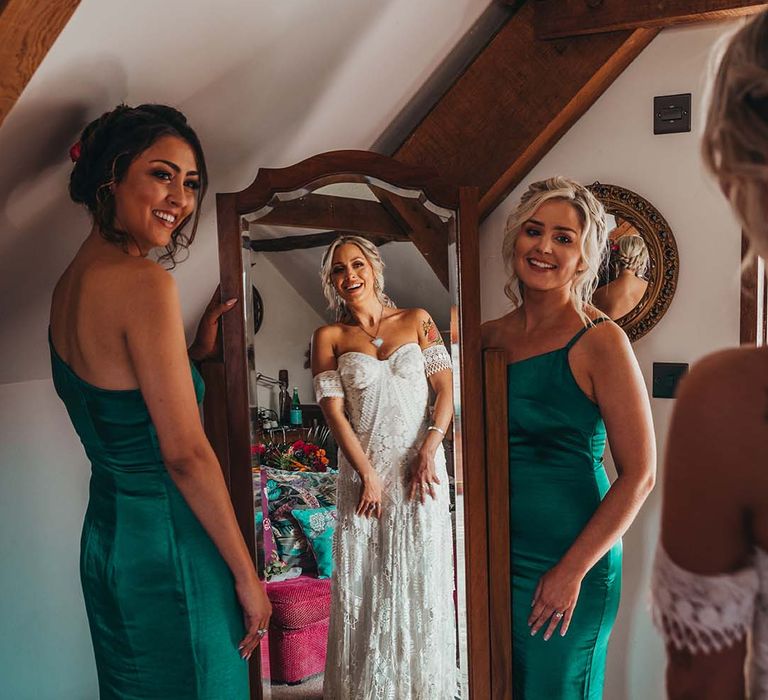 Bride looks at herself in the mirror wearing lace boho wedding dress and bridesmaids in emerald green one shoulder dresses