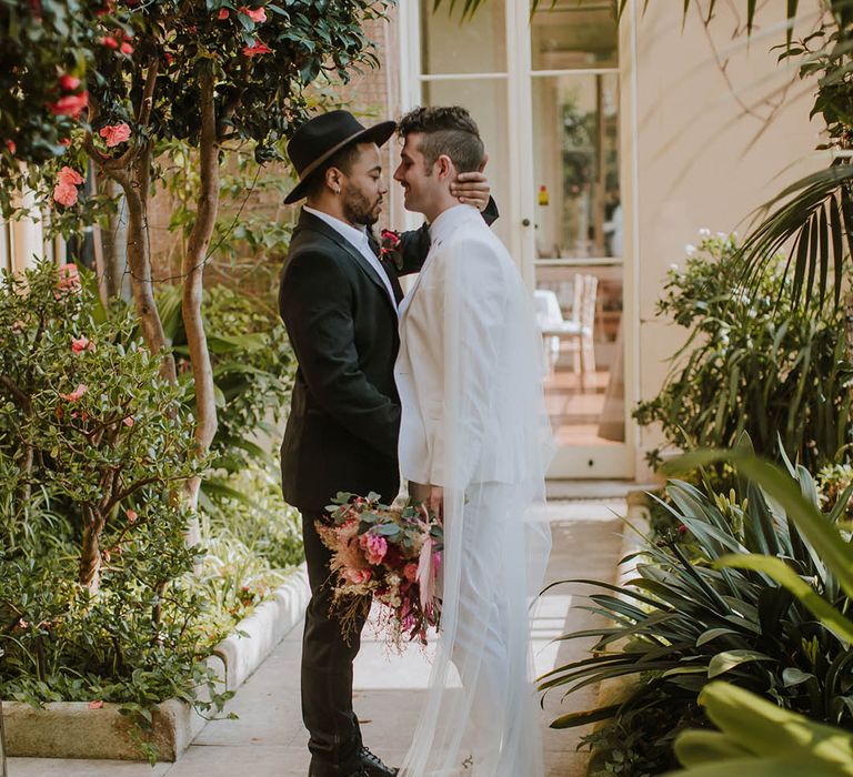 Groom in a black suit and fedora hat embracing his partner in a white groom suit 