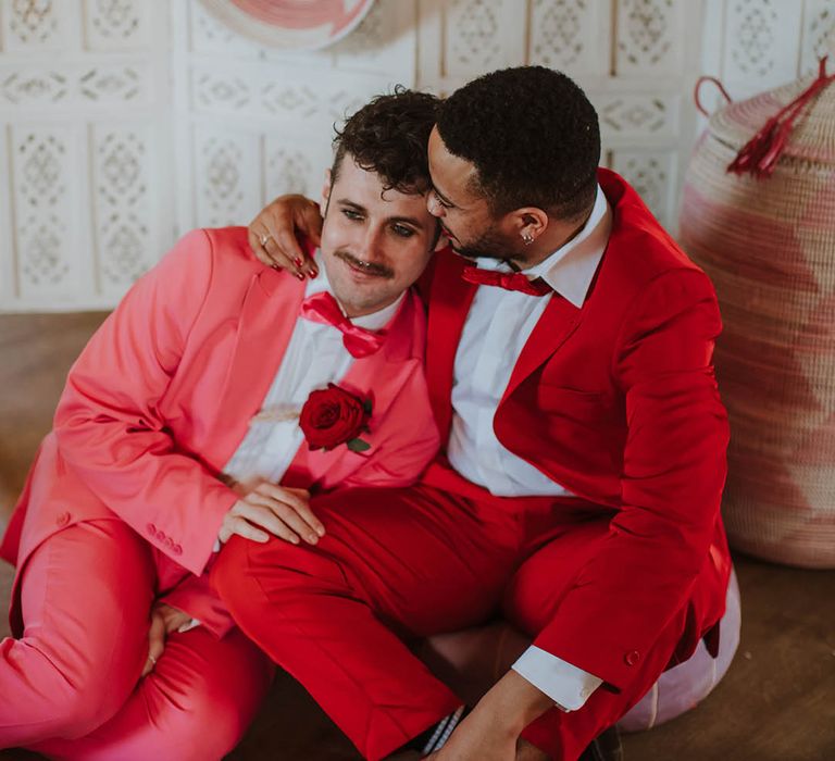 Groom in a red suit with red nail polish embracing his partner on the floor in front of a cream screen and wicker baskets in a pink suit 