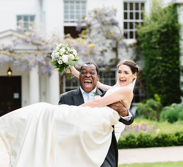 Groom smiles widely as he lifts the bride carrying a purple and white round wedding bouquet 