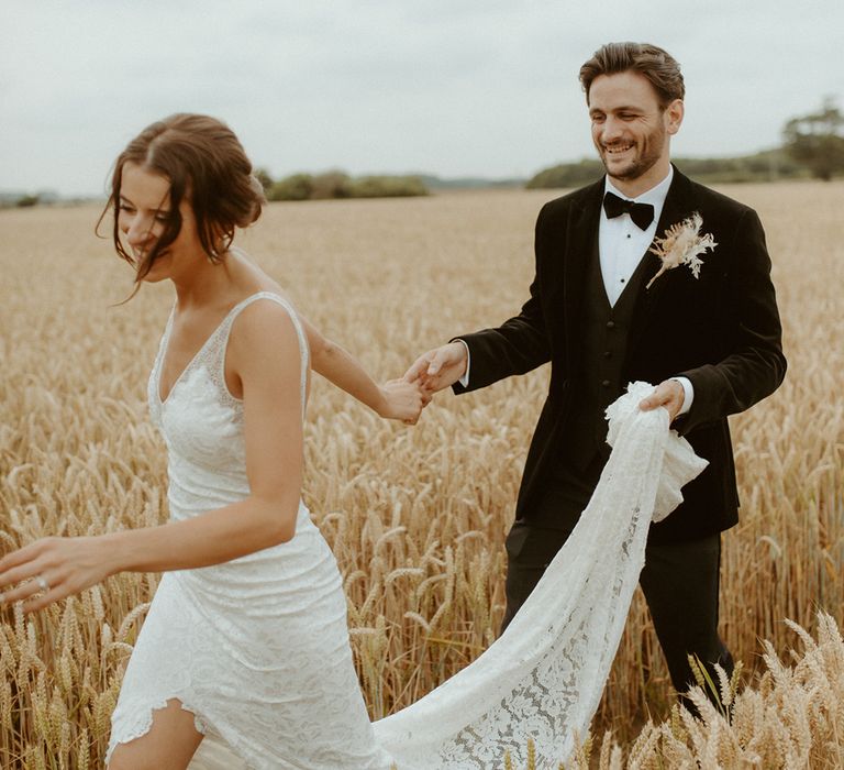 Groom lifts the bride's lace train and holds hands as they walk through a field together for their couple portraits