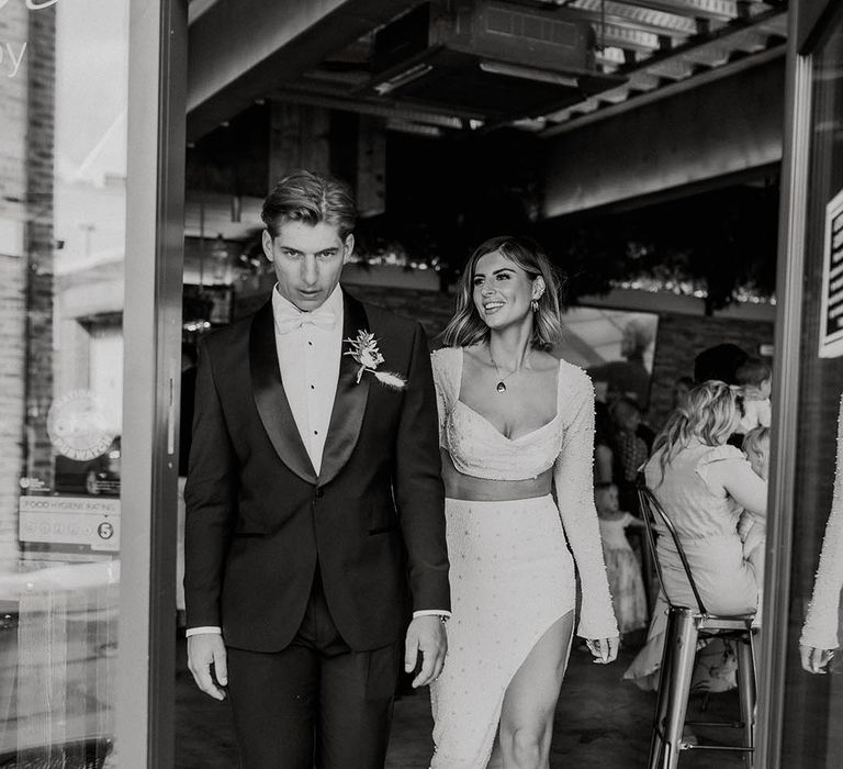 Groom in black suit jacket with satin lapels walking with bride in reception outfit two piece with pearls 