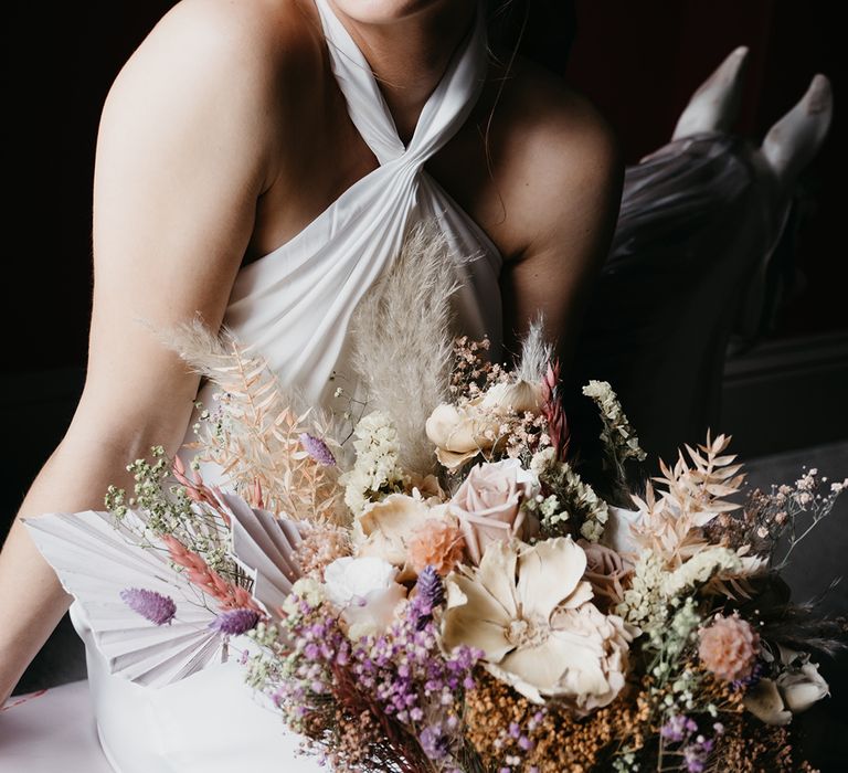 Bride with natural wedding makeup and pinned topknot wearing at satin halter neck wedding dress and preserved pastel flowers and dried gasses wedding bouquet 