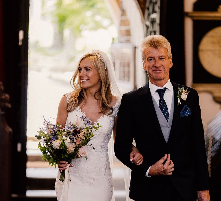 Bride with French tip nails and pearl headband wearing lace wedding dress is walked down the aisle by her father wearing a black and blue suit with polka dots