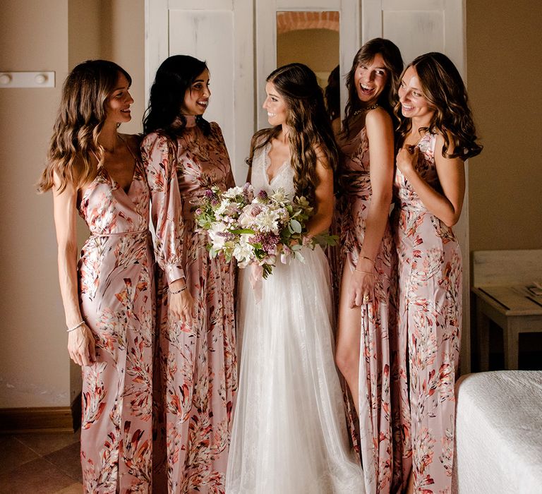 Bride stands with her bridesmaids who wear silk pink floral dresses complete with side leg split 