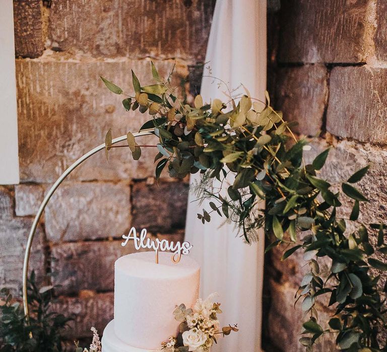 Two tier white wedding cake with textured icing with 'always' cake topper and small circle moongate with flowers