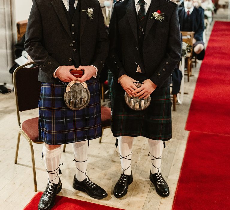 Groom awaits his bride with groomsmen at the front of church and both wear tartan kilts complete with white and red floral buttonholes 