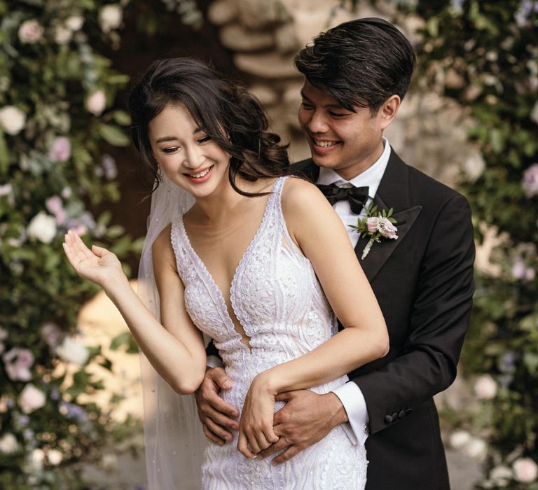 Groom wearing black tie wraps his arms around bride who wears her dark hair in loose curled bun to the side and lace embellished wedding dress 