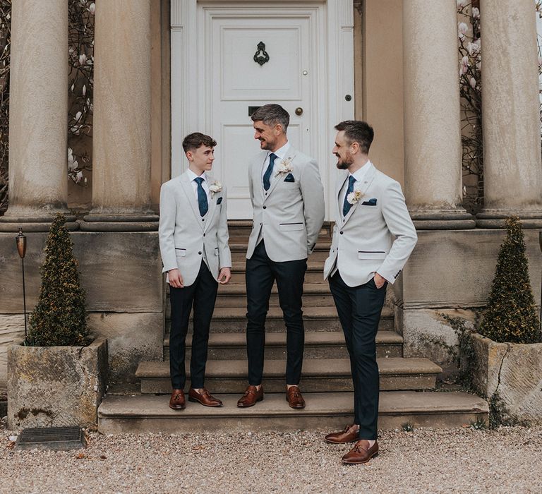 Groom, ring bearer and best man stand together in matching grey suit jackets and blue ties and handkerchiefs and black trousers 