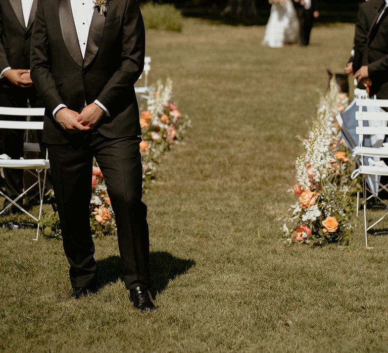 Groom wears black tie suit complete with floral buttonhole as he awaits his bride walking down the aisle 