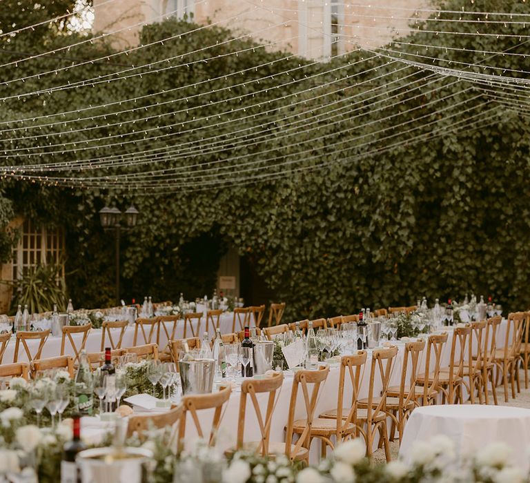 Outdoor wedding reception complete with fairy light canopy hanging above banquet tables finished with white tablecloths and floral table runners 