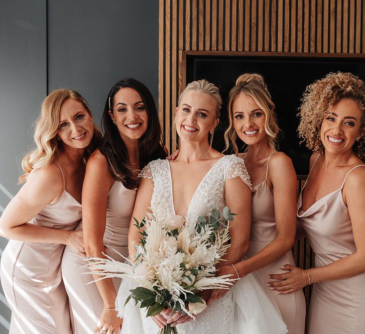 Bride holds neutral bridal bouquet and stands with her bridesmaids wearing silk fitted pastel pink dresses with spaghetti straps and cowl necks