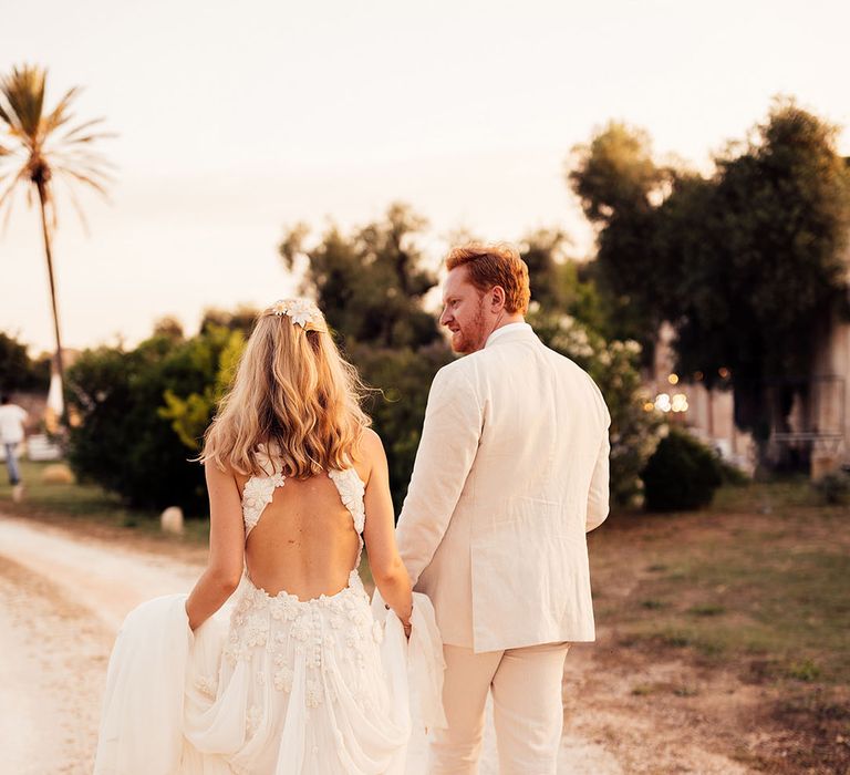 Bride in a flower halter neck wedding dress with flower hair accessories walking with the groom in a cream suit 