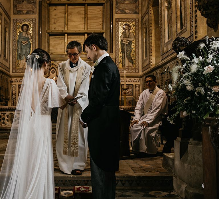 Bride in batwing sleeve and polka dot detail wedding dress and groom in black suit at alter of church wedding with gold accents