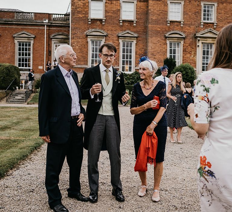 Groom in black suit, sage green waistcoat and tie standing with wedding guests at Kelmarsh Hall country house wedding venue