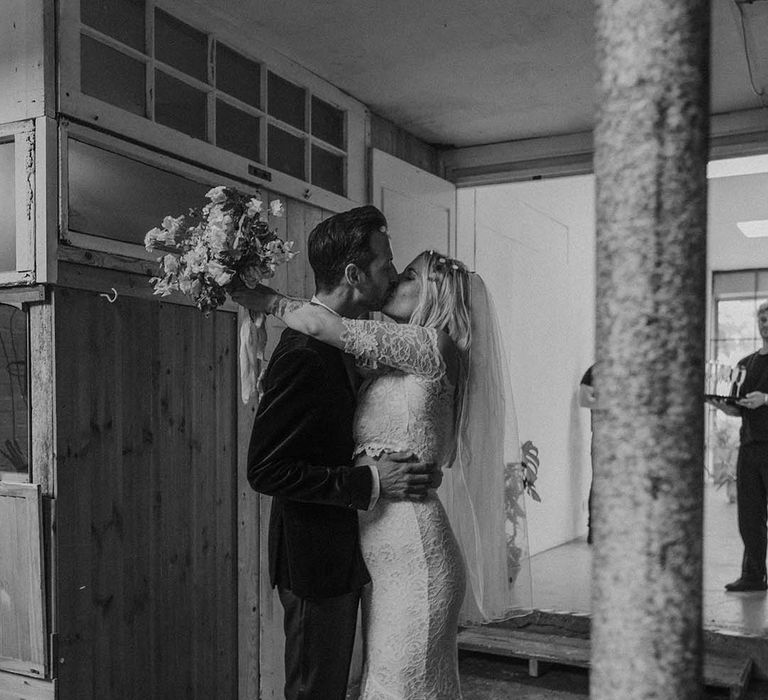 Bride in lace boho two piece with short veil and groom in black tux with bow tie and boutonniere kissing at Hackney Studios