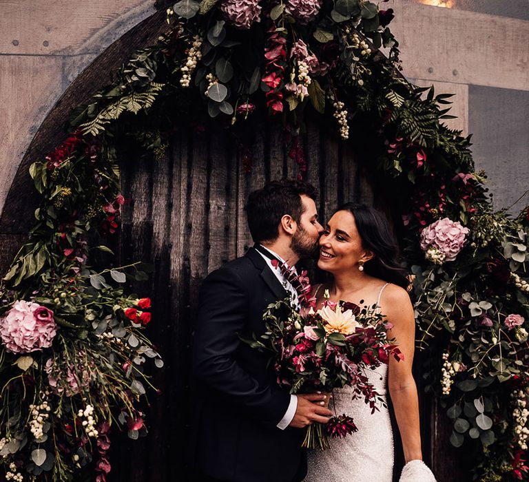 Groom in black tie kisses the bride on the cheek as they stand under a flower arch 