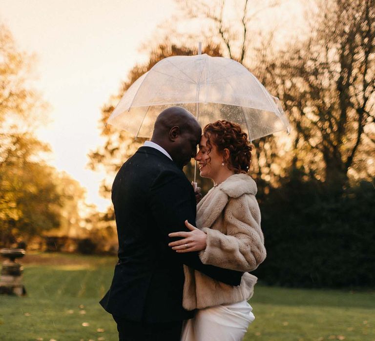 Groom in black tux embracing bride in satin white wedding gown with light fur coat 